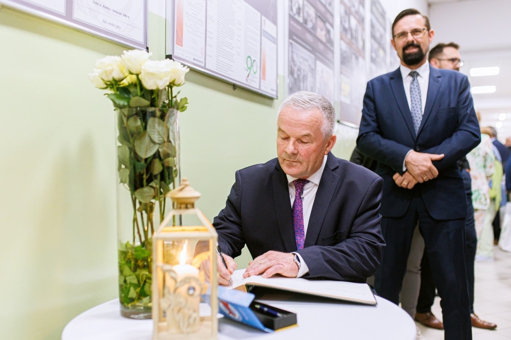 Mężczyzna w garniturze podpisuje dokument przy stole, obok stoi kilka osób, a w tle widoczne są zdjęcia na ścianie.