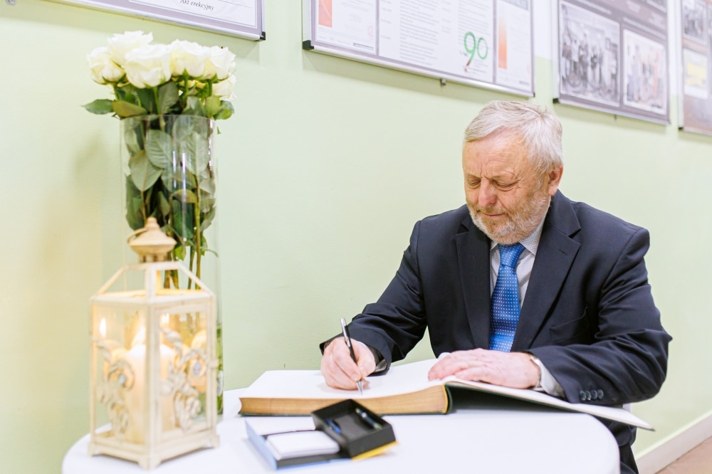 Mężczyzna w garniturze pisze w złotej księdze, obok stoi wazon z białymi różami i lampion.