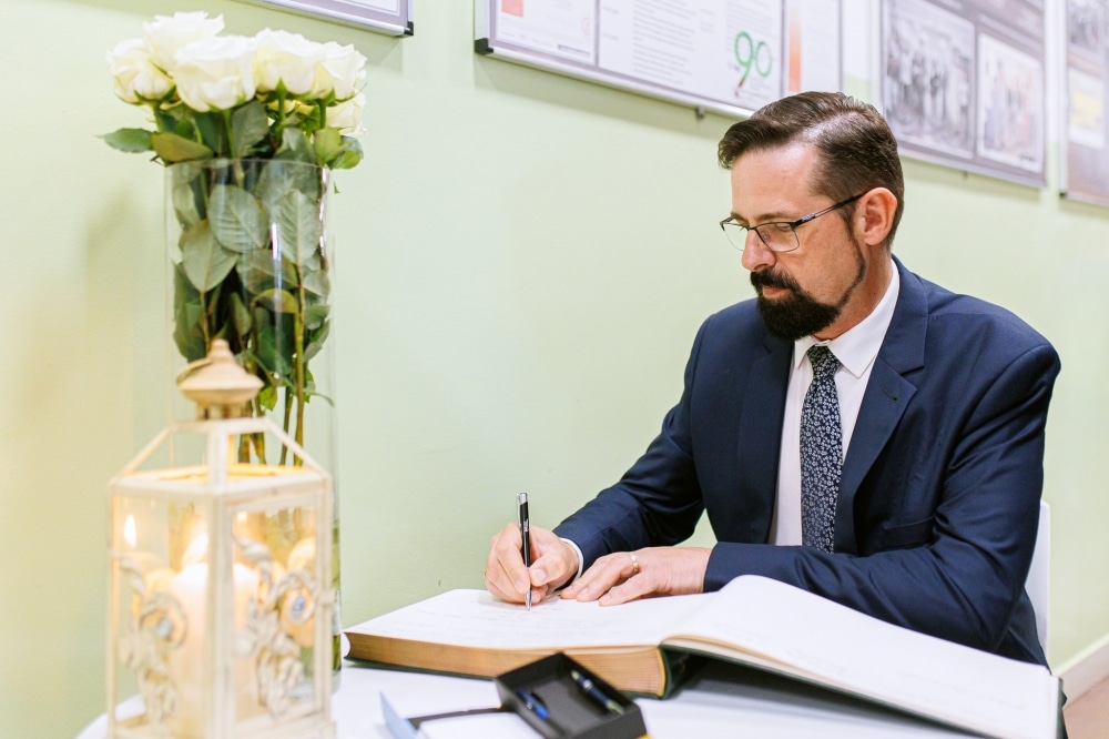 Mężczyzna w eleganckim garniturze pisze w księdze pamiątkowej obok wazonu z białymi różami i latarnią.