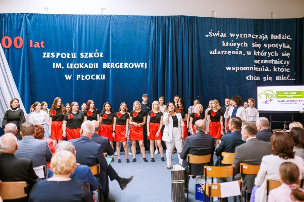 Na zdjęciu widać grupę osób występującą na scenie podczas jubileuszu 100-lecia Zespołu Szkół im. Leokadii Bergerowej w Płocku, z przybyłymi gośćmi w tle.