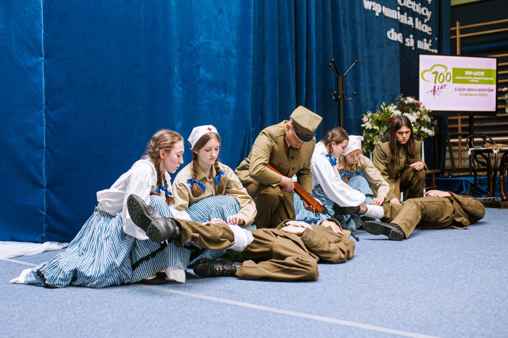 Na zdjęciu przedstawiono grupę młodych ludzi w strojach wojskowych i ludowych, którzy uczestniczą w inscenizacji, w której jeden z nich udziela pomocy medycznej innym.