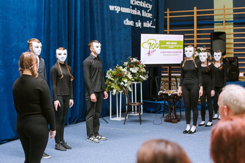 Na zdjęciu widać grupę performerów w czarnych strojach i białych maskach, którzy występują podczas jubileuszu 100-lecia szkoły w Płocku, z niebieskim tłem i dekoracjami w tle.