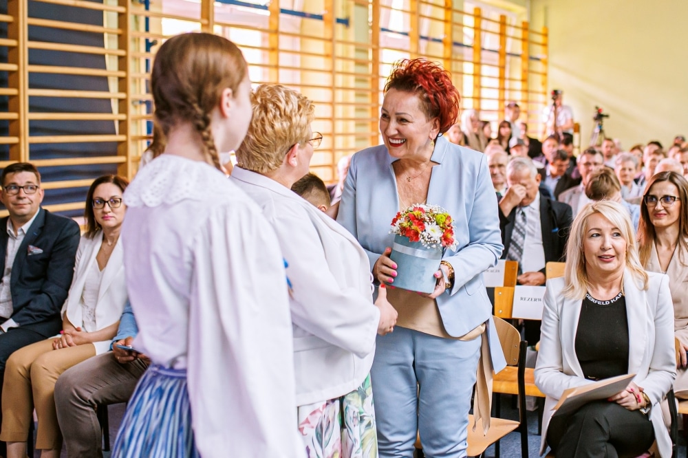 Na zdjęciu widoczna jest uroczystość jubileuszowa, podczas której jedna z kobiet wręcza kwiaty innej, a w tle zgromadzeni goście obserwują wydarzenie.