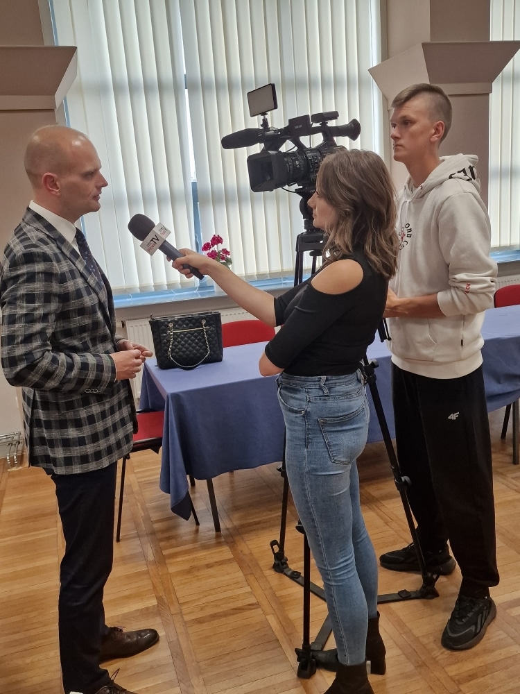 Na zdjęciu dziennikarka przeprowadza wywiad z mężczyzną, podczas gdy operator kamery rejestruje wydarzenie w sali.