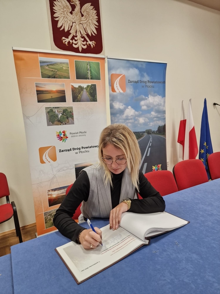 Na zdjęciu osoba siedząca przy stole podpisuje dokumenty w sali konferencyjnej, na tle banerów dotyczących zarządu dróg powiatowych w Płocku.