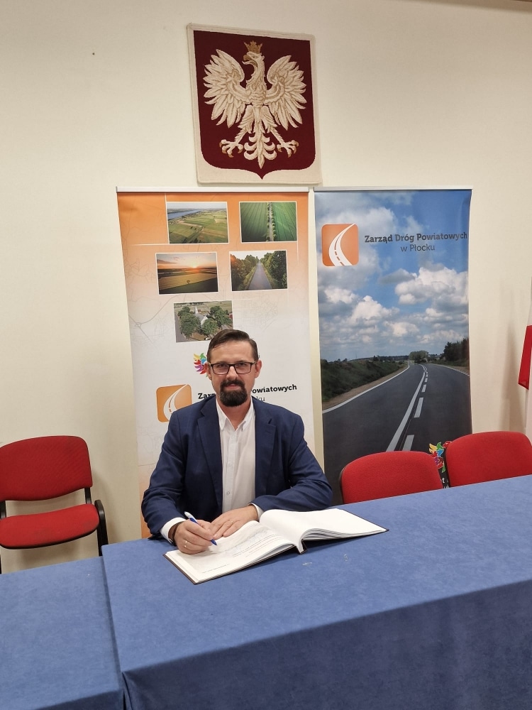 Na zdjęciu widać mężczyznę w garniturze, który podpisuje dokumenty przy stole w sali konferencyjnej, na tle bannerów oraz godła Polski.