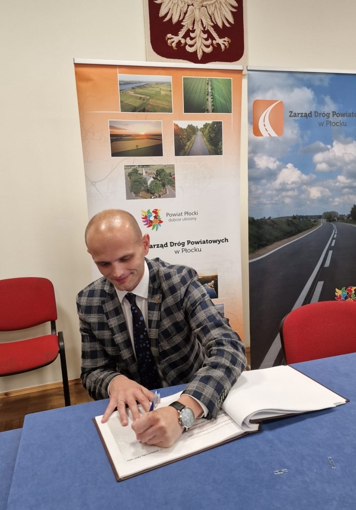 Na zdjęciu widać mężczyznę w eleganckim garniturze, który podpisuje dokumenty przy biurku, w otoczeniu banerów promujących Powiat Płocki.