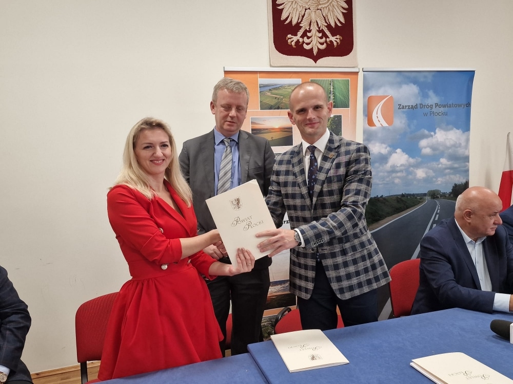 Na zdjęciu widać trzech uczestników wydarzenia, którzy podpisują dokumenty związane z remontem dróg w powiecie płockim, wśród nich kobieta w czerwonej sukience i mężczyzna w wzorzystym garniturze.
