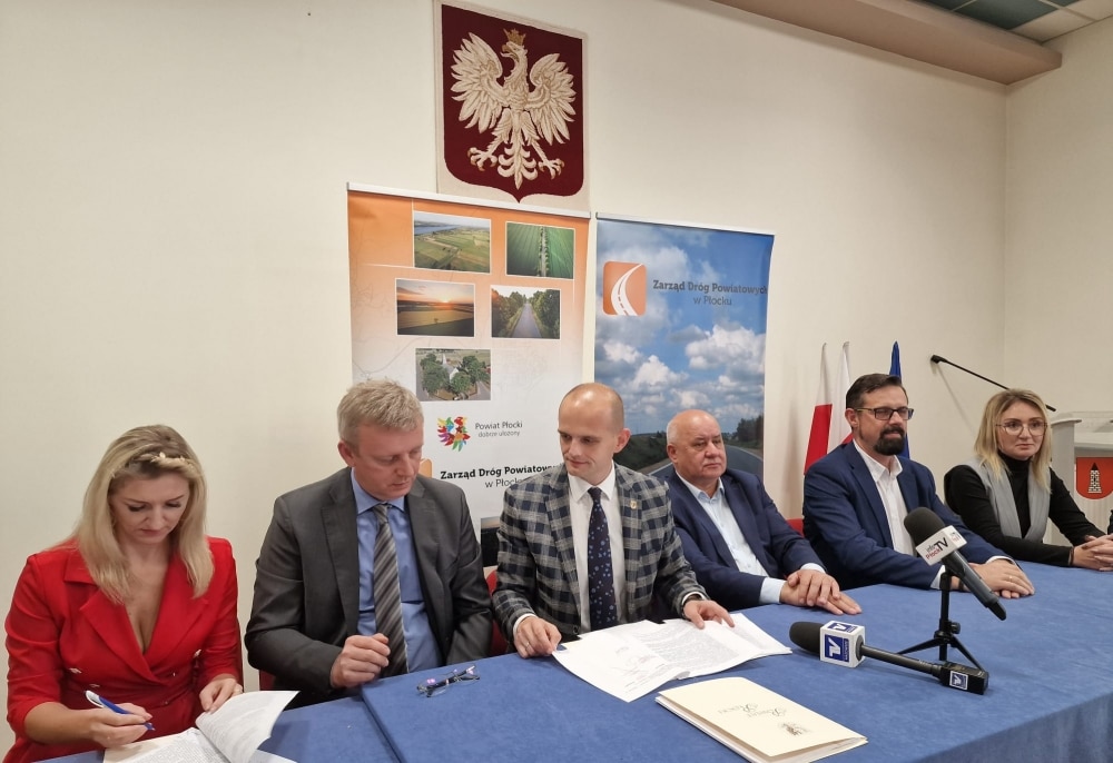 Na zdjęciu widać grupę osób siedzących przy stole podczas konferencji prasowej dotyczącej remontu dróg, z flagami i banerami instytucji lokalnych w tle.