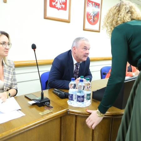 Na zdjęciu przedstawiono posiedzenie rady powiatu, gdzie członkowie dyskutują przy stole, z widocznymi materiałami oraz butelkami z wodą.