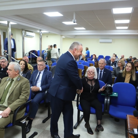 Na zdjęciu widać uczestników sesji Rady Powiatu w Płocku, gdzie jeden z mężczyzn witającego się z kobietą w okularach, a w tle siedzi grupa osób w niebieskich fotelach.