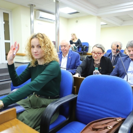 Na zdjęciu widać osoby uczestniczące w sesji rady powiatu, w tym kobietę z kręconymi włosami, która unosi rękę.