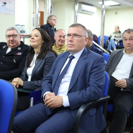 Na zdjęciu przedstawiono grupę osób siedzących na spotkaniu, w tym mężczyznę w okularach oraz kobietę w jasnej bluzce, w sali konferencyjnej z niebieskimi krzesłami.