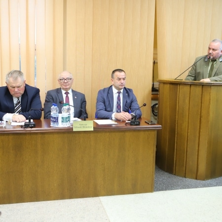 Na zdjęciu przedstawiono posiedzenie rady powiatu, gdzie czterech mężczyzn siedzi przy stole, a jeden z nich przemawia z podium.