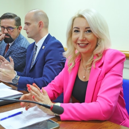 Na zdjęciu widać uśmiechającą się kobietę w różowej marynarce siedzącą przy stole podczas spotkania, obok niej dwóch mężczyzn w garniturach.