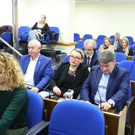 Na zdjęciu widoczna jest grupa ludzi siedzących w sali obrad, z zamontowanymi mikrofonami i niebieskimi krzesłami, w czasie sesji rady powiatu.