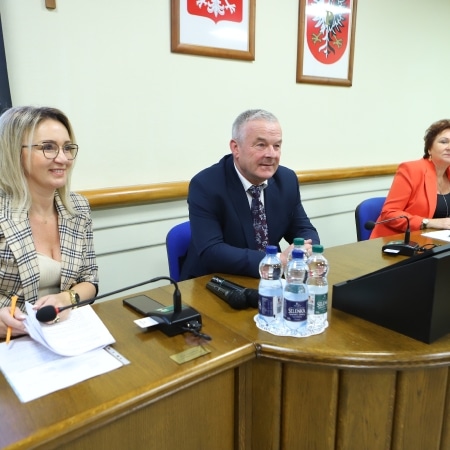 Na zdjęciu widoczna jest trójka ludzi podczas spotkania, z zaangażowaniem rozmawiających przy stole w sali konferencyjnej.