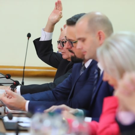 Na zdjęciu widać członków rady powiatu, którzy biorą udział w posiedzeniu, podnosząc rękę w geście głosowania.