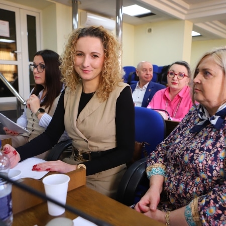 Na zdjęciu znajduje się grupa osób podczas sesji Rady Powiatu w Płocku, w której jedna z kobiet w beżowym ubraniu uśmiecha się do aparatu, a w tle siedzą inni uczestnicy.