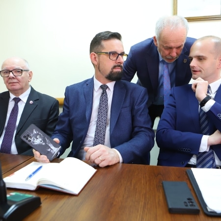 Na zdjęciu widać grupę mężczyzn w garniturach, którzy siedzą przy stole w sali konferencyjnej, z zaangażowaniem dyskutując na temat dokumentów i telefonów.