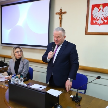 Na zdjęciu widoczna jest sala konferencyjna, w której mężczyzna w garniturze przemawia do zebranych, a obok niego siedzi uśmiechnięta kobieta w okularach.