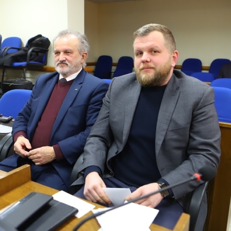 Na zdjęciu widoczni są dwaj mężczyźni siedzący w sali obrad, z poważnymi wyrazami twarzy.