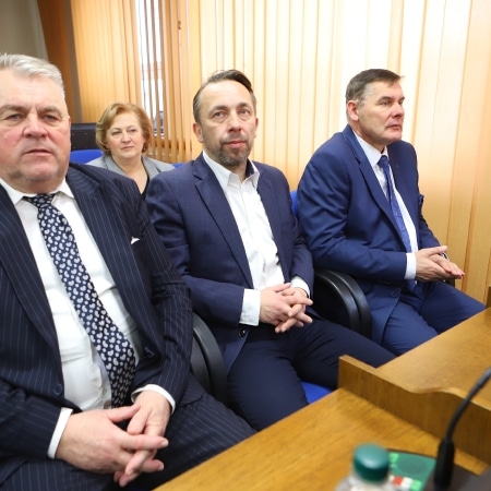 Na zdjęciu widoczna jest czwórka ludzi, z których trzech mężczyzn siedzi przy stole, a jedna kobieta stoi za nimi, w tle widać jasne zasłony.