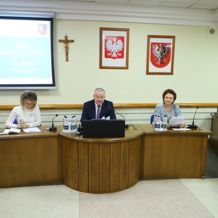 Na zdjęciu przedstawiono sesję Rady Powiatu w Płocku, podczas której dyskutowane są ważne kwestie, a wśród uczestników są trzy osoby przy stole prezydialnym.