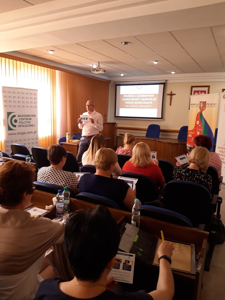 Zdjęcie przedstawia grupę uczestników szkolenia, w trakcie którego omawiane są problemy uzależnień i dysfunkcji relacji społecznych, a prowadzący prezentuje materiały edukacyjne.