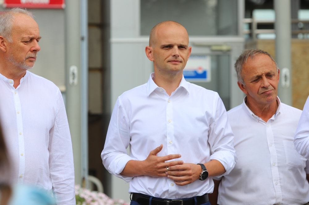 Na zdjęciu widoczna jest grupa trzech mężczyzn w białych koszulach, z poważnymi wyrazami twarzy, stojących w publicznym miejscu.