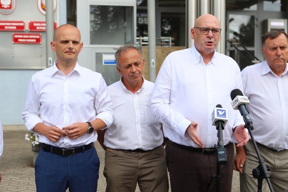 Na zdjęciu widoczna jest grupa mężczyzn w białych koszulach, którzy prowadzą konferencję prasową w plenerze, wyrażając swoje zaniepokojenie sprawami rolników.