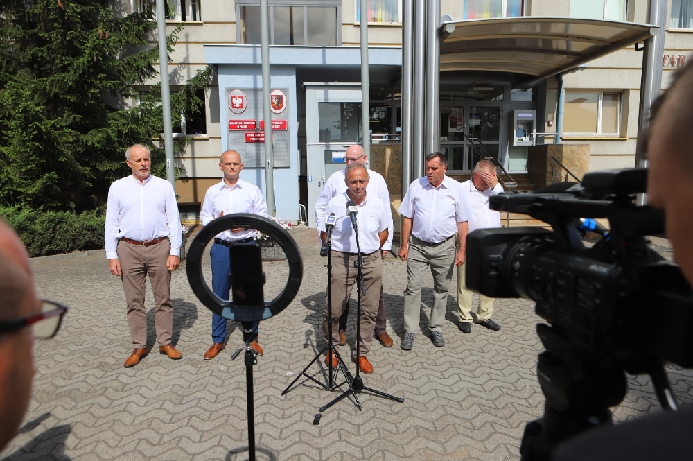 Grupa mężczyzn w białych koszulach stoi przed budynkiem, prowadząc konferencję prasową w sprawie sytuacji rolników.