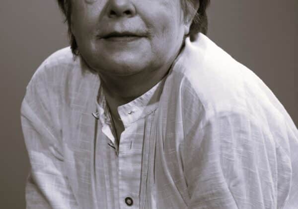 Portret starszej kobiety z krótkimi włosami, ubranej w jasną bluzkę, z wyrazem twarzy pełnym refleksji, siedzącej z założonymi rękami.
