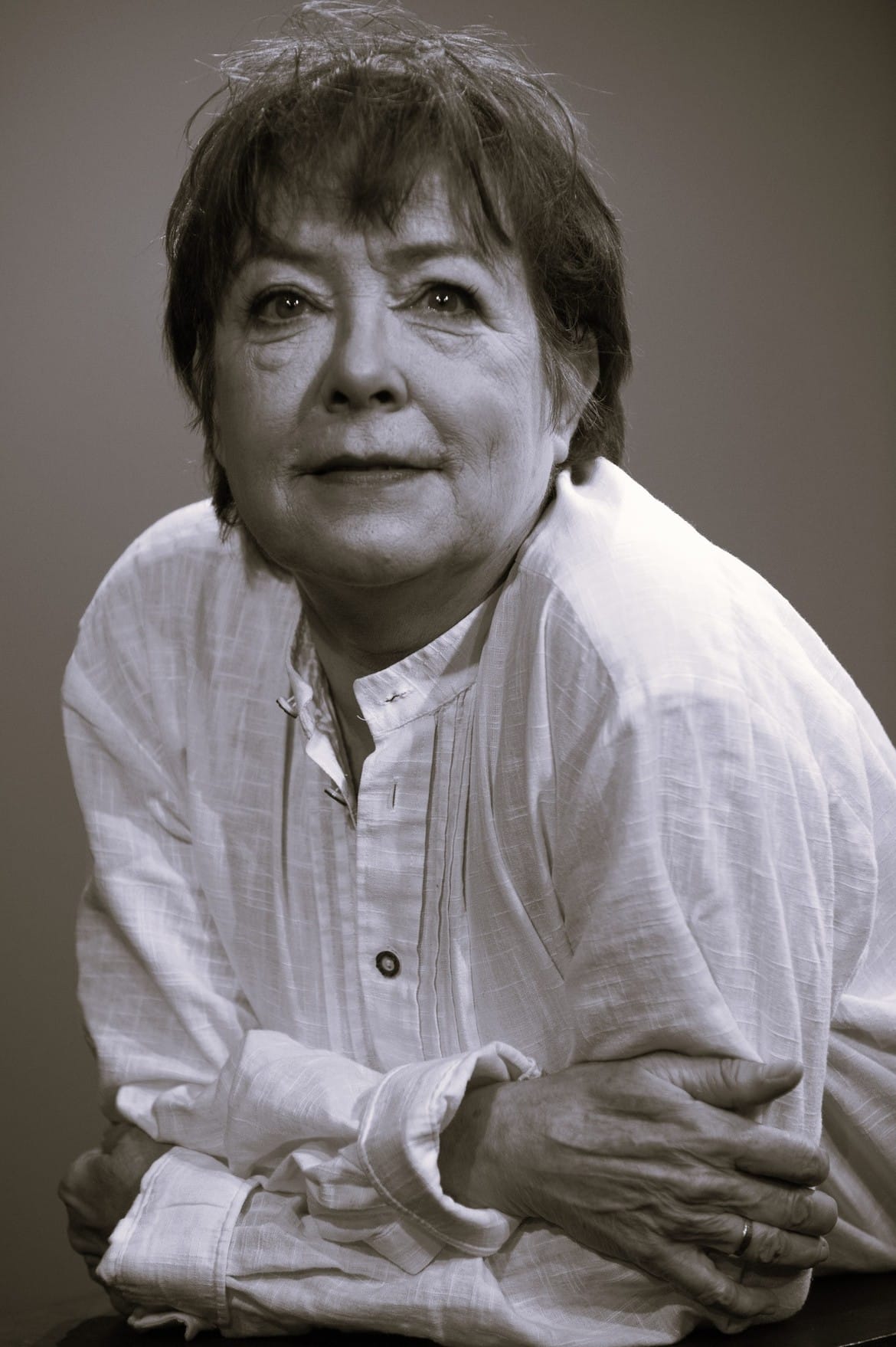 Portret starszej kobiety z krótkimi włosami, ubranej w jasną bluzkę, z wyrazem twarzy pełnym refleksji, siedzącej z założonymi rękami.