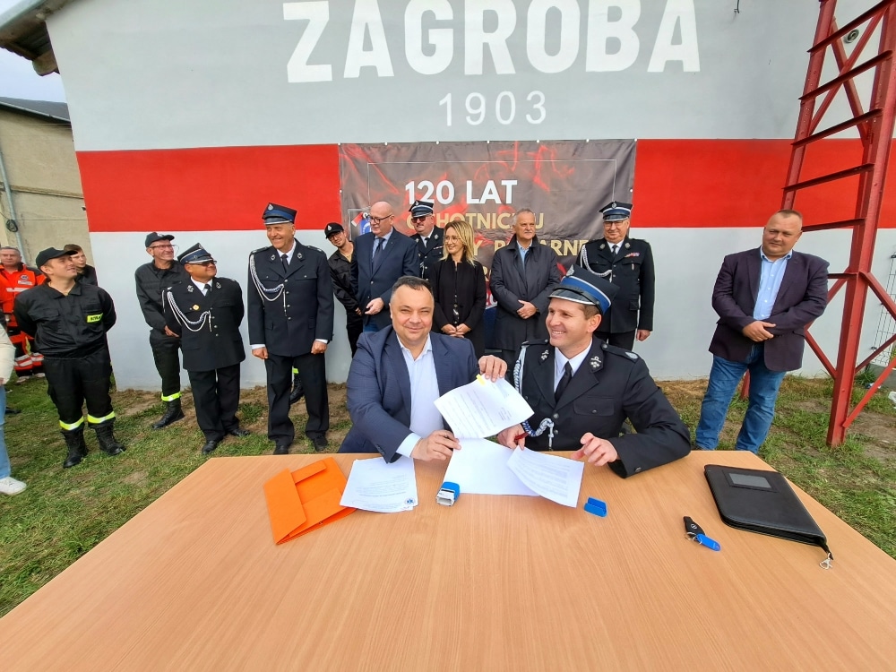 Na zdjęciu przedstawiono dwóch mężczyzn podpisujących umowę przy stole, podczas uroczystości z okazji 120-lecia Ochotniczej Straży Pożarnej w Zagrobie, z grupą strażaków i gości w tle.