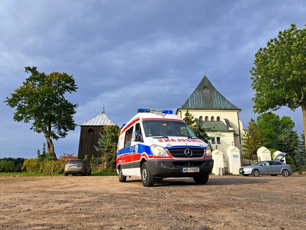 Na zdjęciu widać ambulans zaparkowany na terenie w pobliżu kościoła w malowniczej okolicy, otoczonej drzewami i chmurami na niebie.