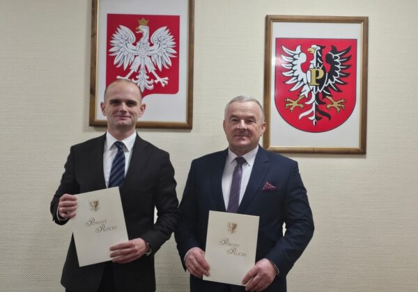 Na zdjęciu dwóch mężczyzn w garniturach trzymających dokumenty, stojących w biurze, z godłami Polski i powiatu w tle, informuje o rekompensacie zniszczeń na drogach powiatowych przez Orlen.