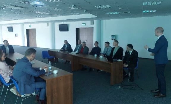 Zdjęcie przedstawia spotkanie grupy mężczyzn i jednej kobiety przy długim stole w nowoczesnym pomieszczeniu, gdzie ktoś prowadzi prezentację.