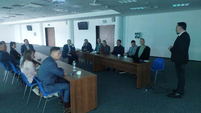 Na zdjęciu przedstawiono spotkanie grupy ludzi w sali konferencyjnej, podczas którego jeden z uczestników prowadzi prezentację.