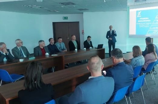 Na zdjęciu przedstawiono grupę osób uczestniczących w spotkaniu w sali konferencyjnej, z mężczyzną wygłaszającym wykład na tle prezentacji.