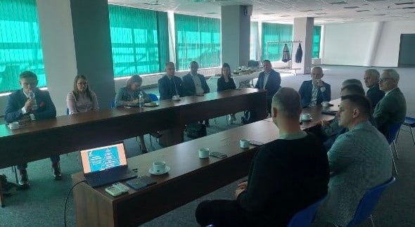 Zdjęcie przedstawia grupę osób siedzących przy długim stole w sali konferencyjnej podczas spotkania, z ekranem projekcyjnym i filiżankami na stole.