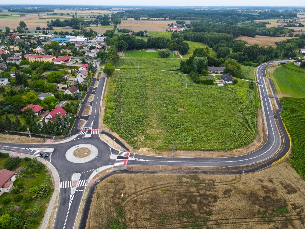 Zdjęcie przedstawia nowo wybudowane rondo z otaczającą infrastrukturą drogową oraz sąsiednią zabudową mieszkalną i terenami zielonymi w okolicy wiejskiej.