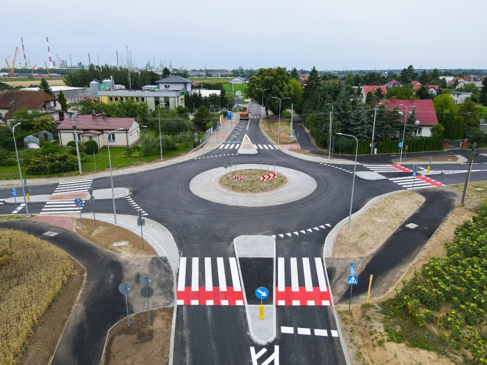 Zdjęcie przedstawia nowo zbudowane rondo w otoczeniu domów i zieleni, z wyraźnie oznakowanymi pasami i znakami drogowymi.