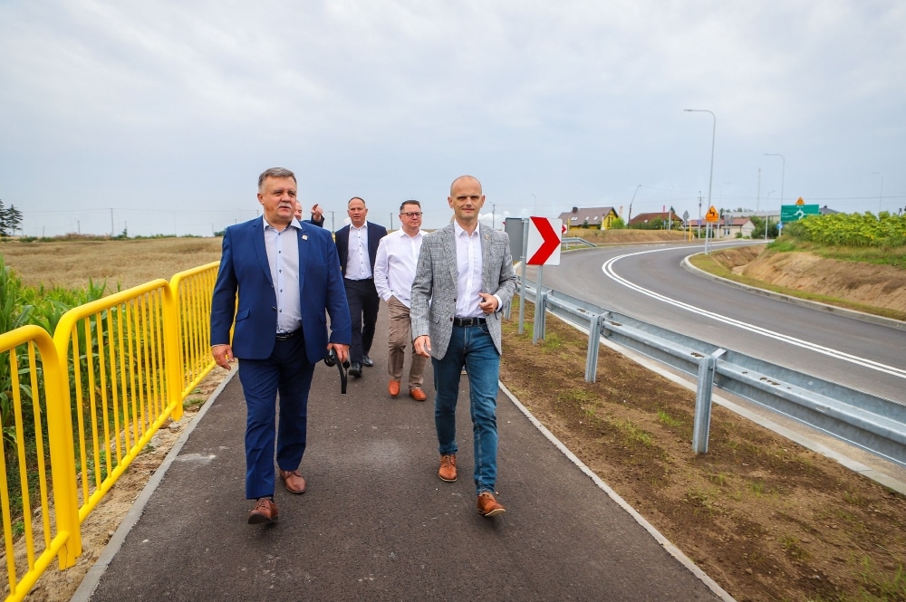 Na zdjęciu grupa mężczyzn w garniturach spaceruje po nowo wybudowanej drodze obok ronda.