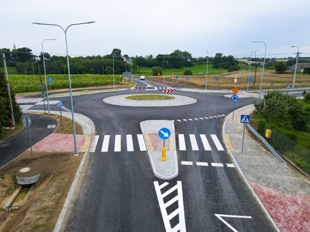 Nowoczesne rondo z oznakowaniem drogowym i ścieżkami dla pieszych, otoczone zielenią.