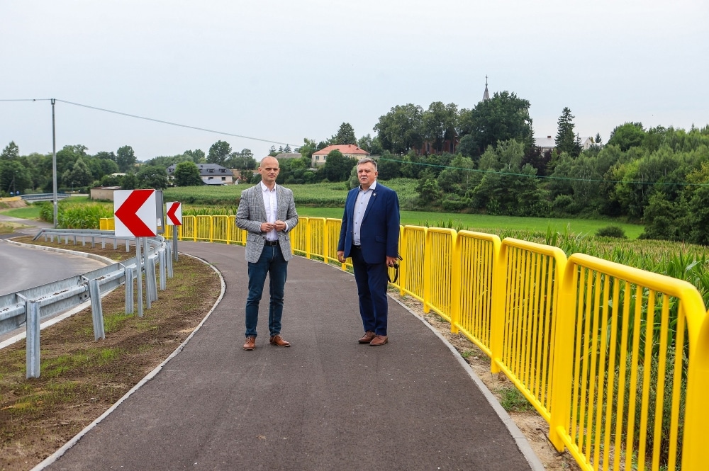 Na zdjęciu widać dwóch mężczyzn stojących przy nowo otwartym rondzie, otoczonym zielenią i żółtymi barierkami.