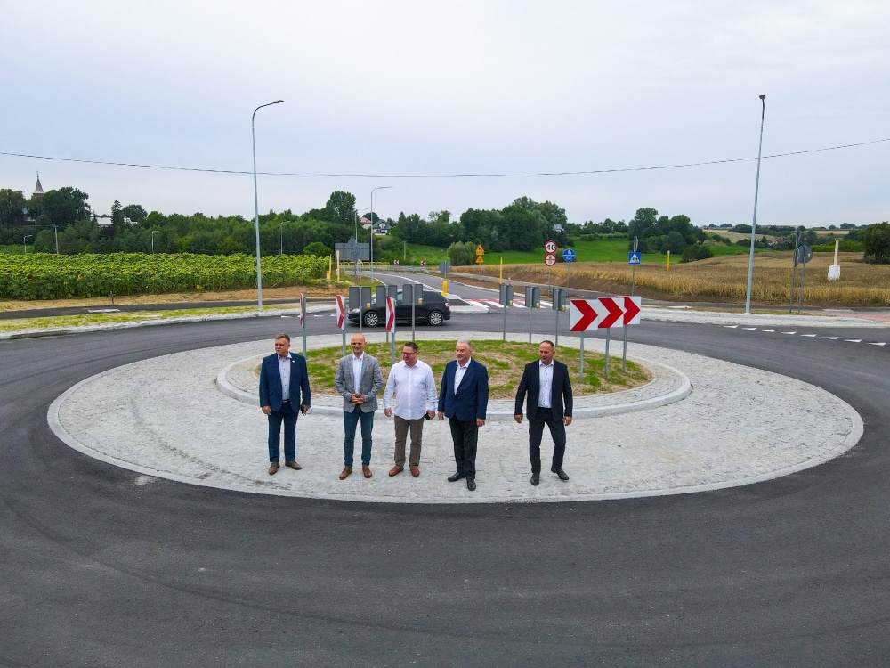 Na zdjęciu widoczna jest nowo wybudowana rondo z grupą mężczyzn stojących na jego środku w otoczeniu zieleni i znaków drogowych.