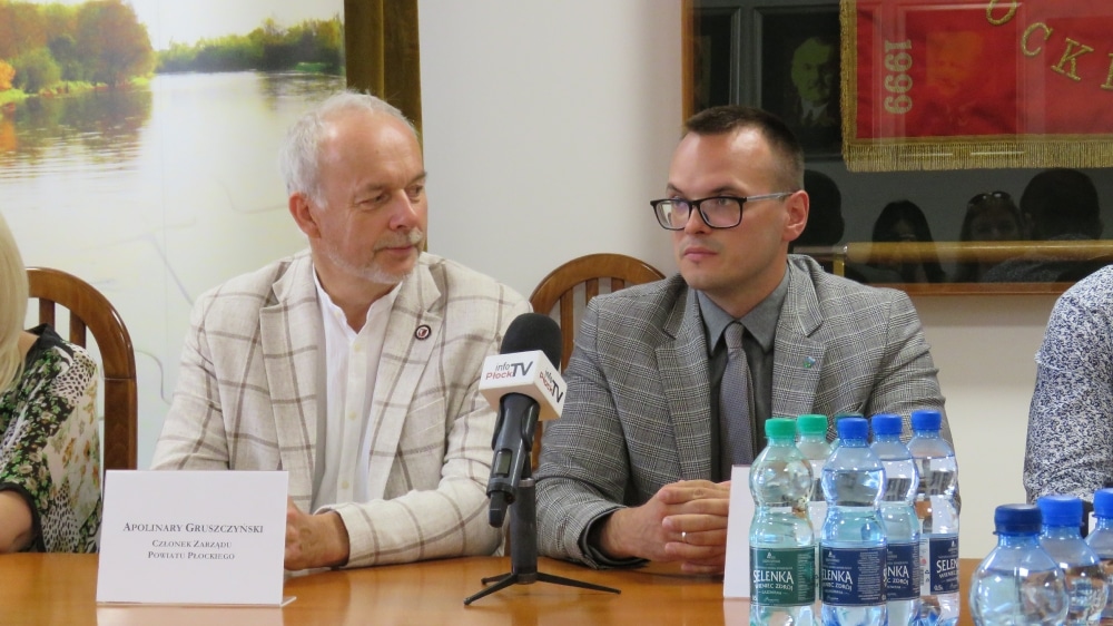 Na zdjęciu przedstawieni są mężczyźni podczas konferencji prasowej, z wodą butelkowaną na stole oraz tłem z krajobrazem.