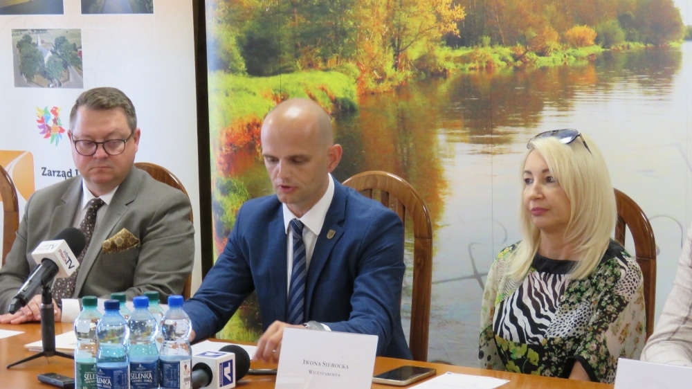 Na zdjęciu widoczna jest trójka osób podczas konferencji prasowej, z tłem przedstawiającym krajobraz nad wodą.