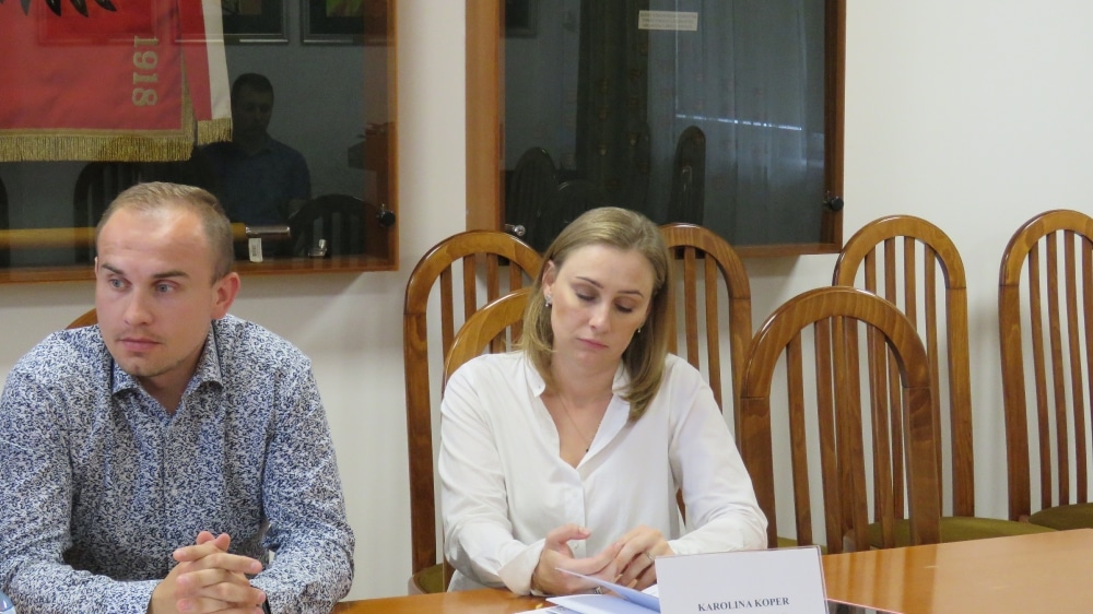 Na zdjęciu przedstawione są dwie osoby siedzące przy stole w sali konferencyjnej, z widocznym tłem i flagą.